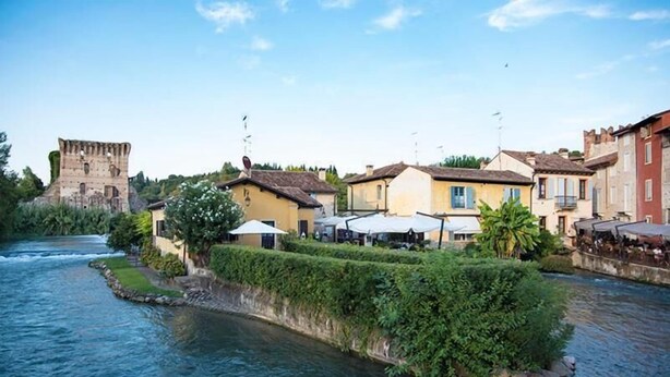 A riverside town with historic architecture, including a castle-like structure, and buildings with outdoor seating areas. - Le Finestre su Borghetto (Valeggio sul Mincio)