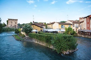 A riverside town with historic architecture, including a castle-like structure, and buildings with outdoor seating areas. - Le Finestre su Borghetto (Valeggio sul Mincio)