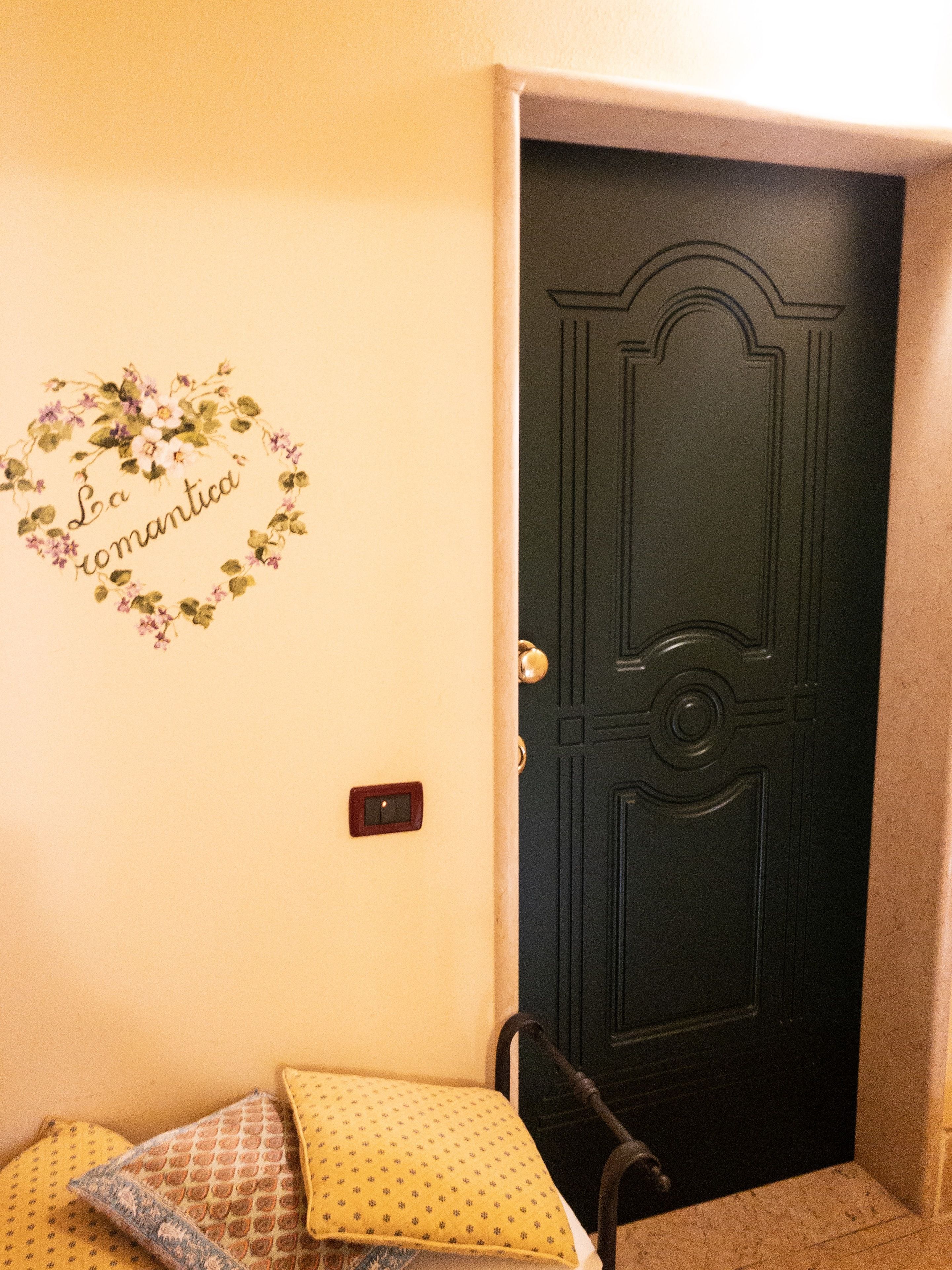 Une pièce avec une porte sombre, une décoration murale portant l’inscription « La romantica », et un lit agrémenté d’oreillers à motifs.