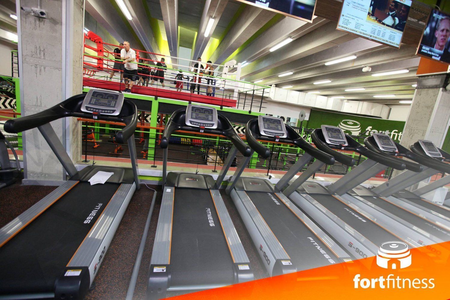 A gym with multiple treadmills, a boxing ring, and a green wall with a logo.