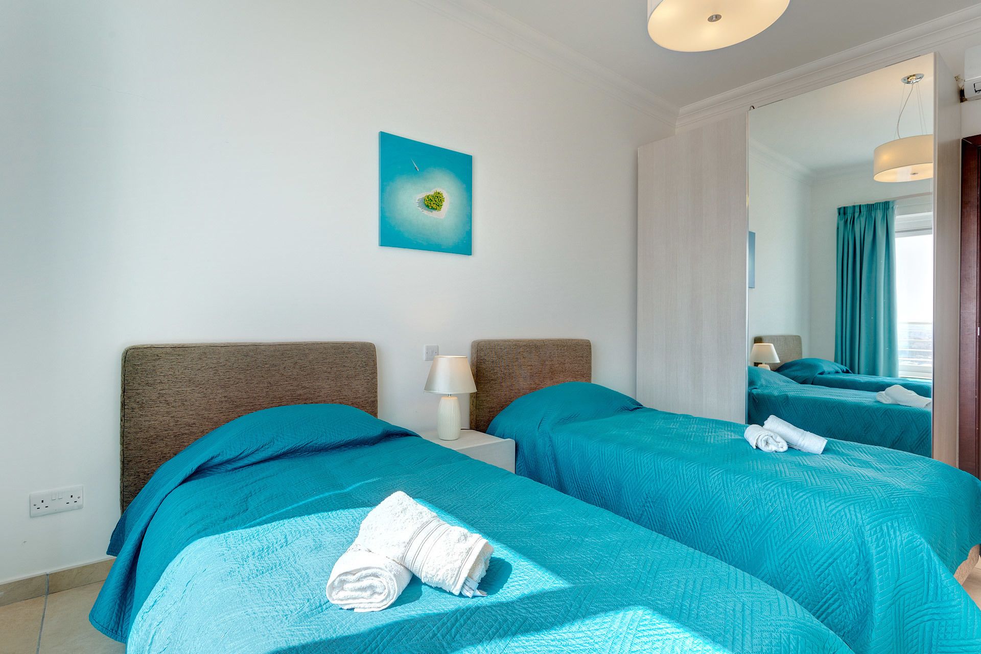 A hotel room with two beds, a painting on the wall, and a mirror reflecting the room.