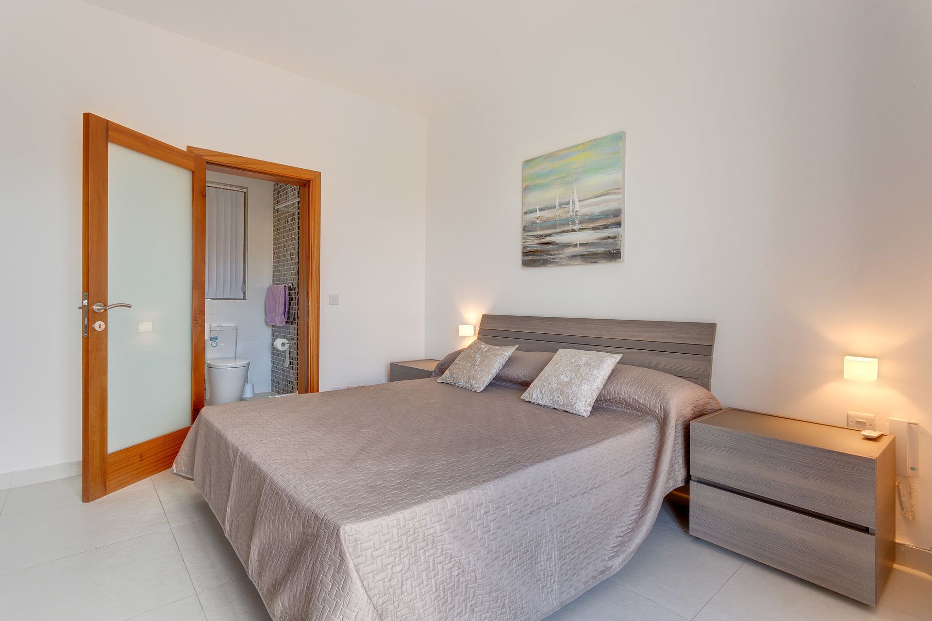 A bedroom with a bed, bedside tables, a nightstand, a painting on the wall, and a bathroom visible through an open door.