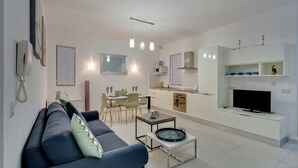Un salon moderne avec un canapé, une table basse et un téléviseur. Il y a également un coin repas avec des chaises et une table. La cuisine est dotée de meubles blancs et d’un téléviseur.