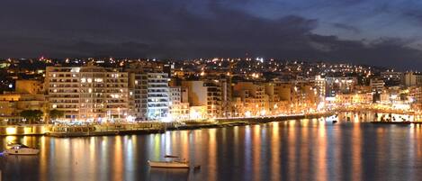 A nighttime cityscape with buildings along a waterfront, boats in the water, and a calm river reflecting the lights.