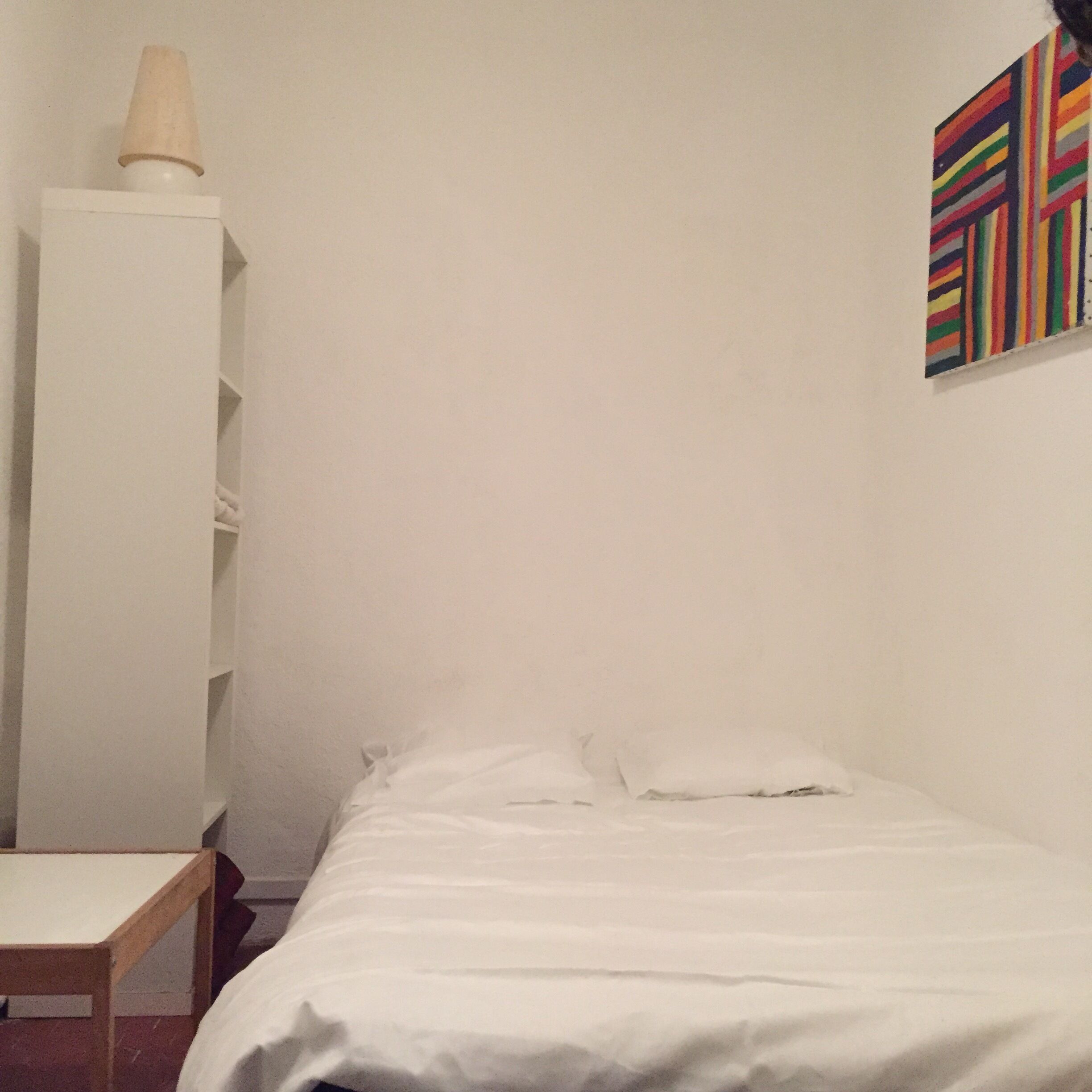 Une pièce simple, avec un lit, une armoire blanche et un tableau abstrait aux couleurs vives accroché au mur.