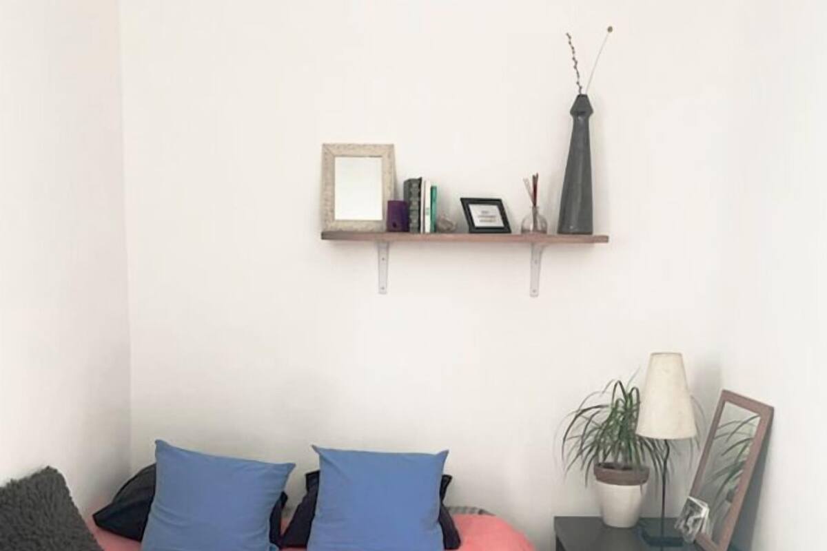 Une chambre à coucher comprenant un lit, des oreillers, une table de chevet, une lampe, une petite table et une étagère murale décorée de divers objets.