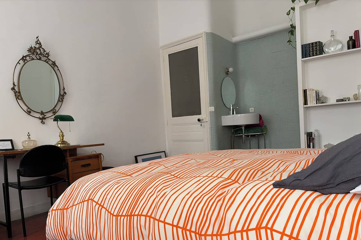 Une chambre à coucher comprenant un lit, un bureau avec une chaise, une commode, un miroir, une étagère avec des livres et des objets décoratifs, ainsi qu’une salle de bain avec un lavabo et un miroir.
