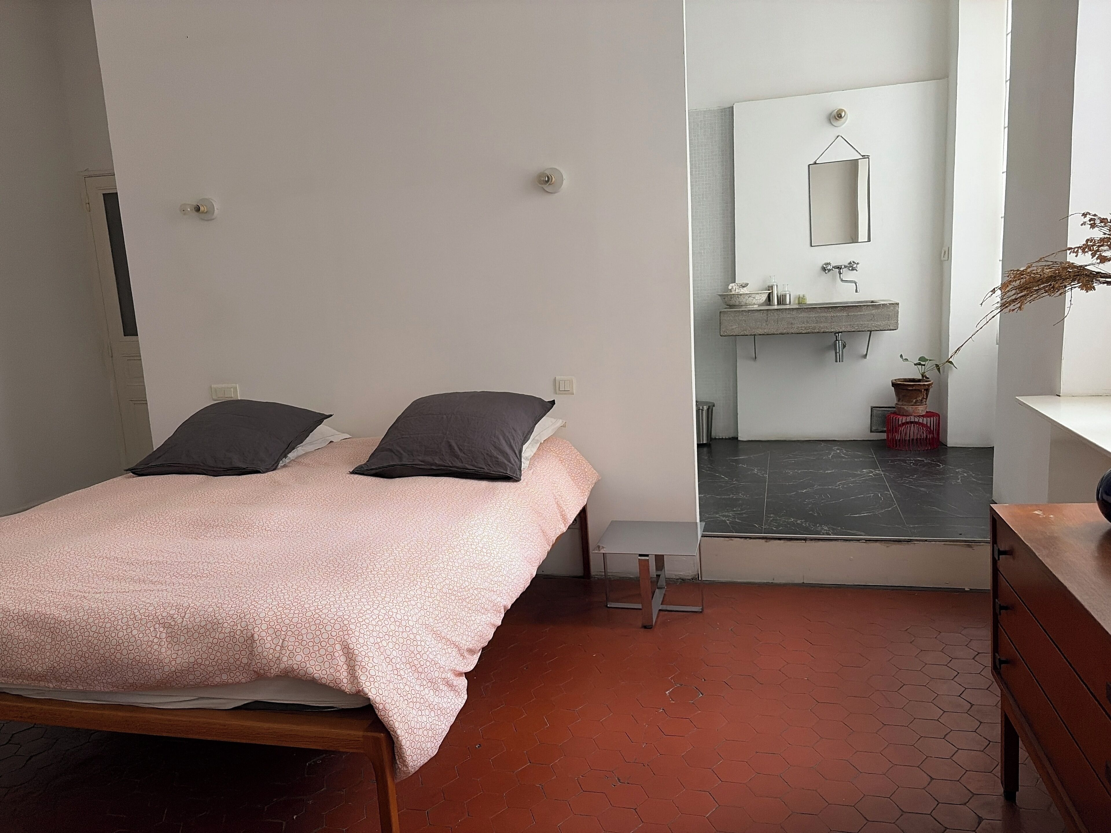 Un dormitorio con una cama, una mesita de noche, un espejo y un baño visible a través de una puerta abierta.