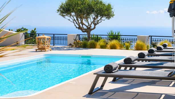 A swimming pool with lounge chairs, a tree, and a view of the sea. - Grand Villa Santorini (Santorini)