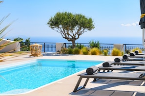 A swimming pool with lounge chairs, a tree, and a view of the sea. - Grand Villa Santorini (Santorini)