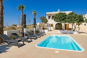 A swimming pool with lounge chairs, palm trees, and a white building with a balcony. - Grand Villa Santorini (Santorini)