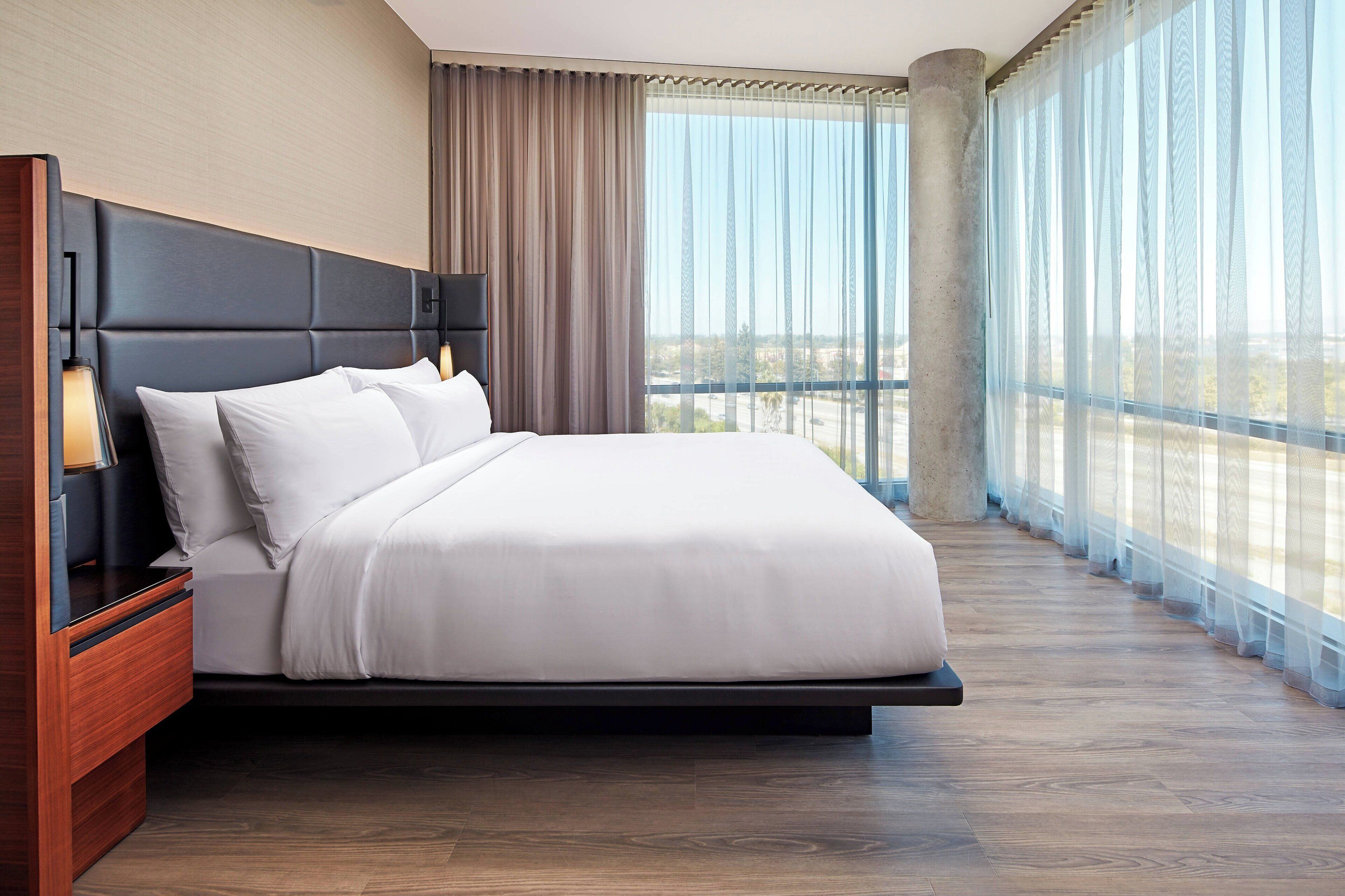 Una habitación de hotel moderna con una cama grande, una mesita de noche de madera y vistas a la ciudad a través de amplios ventanales.