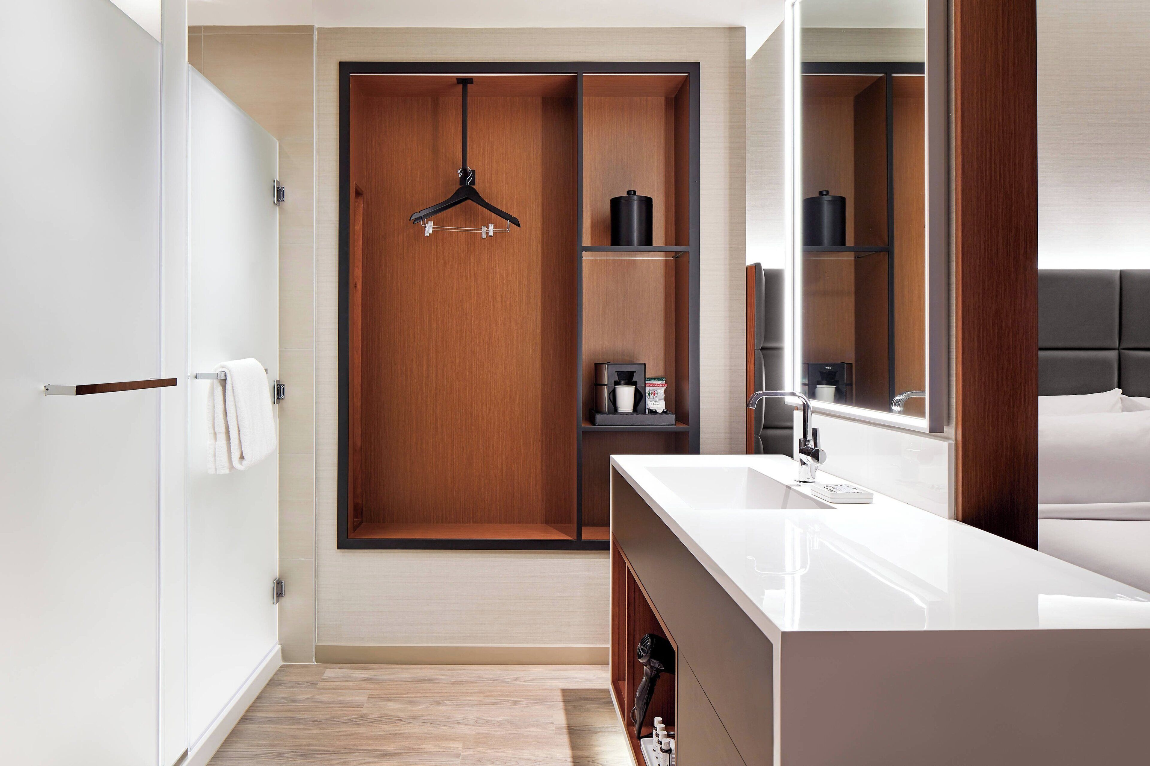 A modern hotel bathroom with a wooden vanity, a large mirror, and a walk-in closet with a hanging rack.