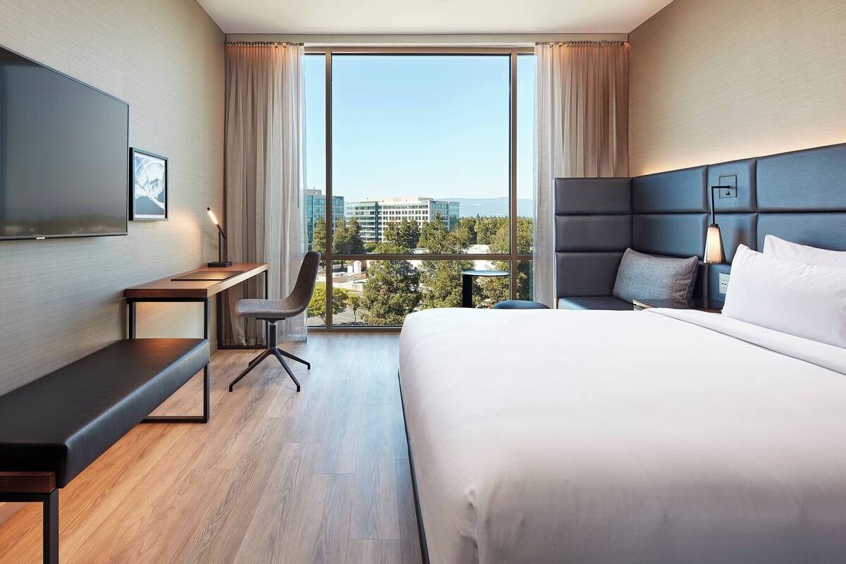 Ein modernes Hotelzimmer mit einem großen Bett, einem Schreibtisch, einem Sessel, einem Fernseher und Blick auf die Stadt durch große Fenster.