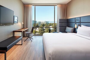 Habitación de hotel moderna con una cama grande, un escritorio, una silla, un televisor y vistas de la ciudad a través de amplias ventanas.