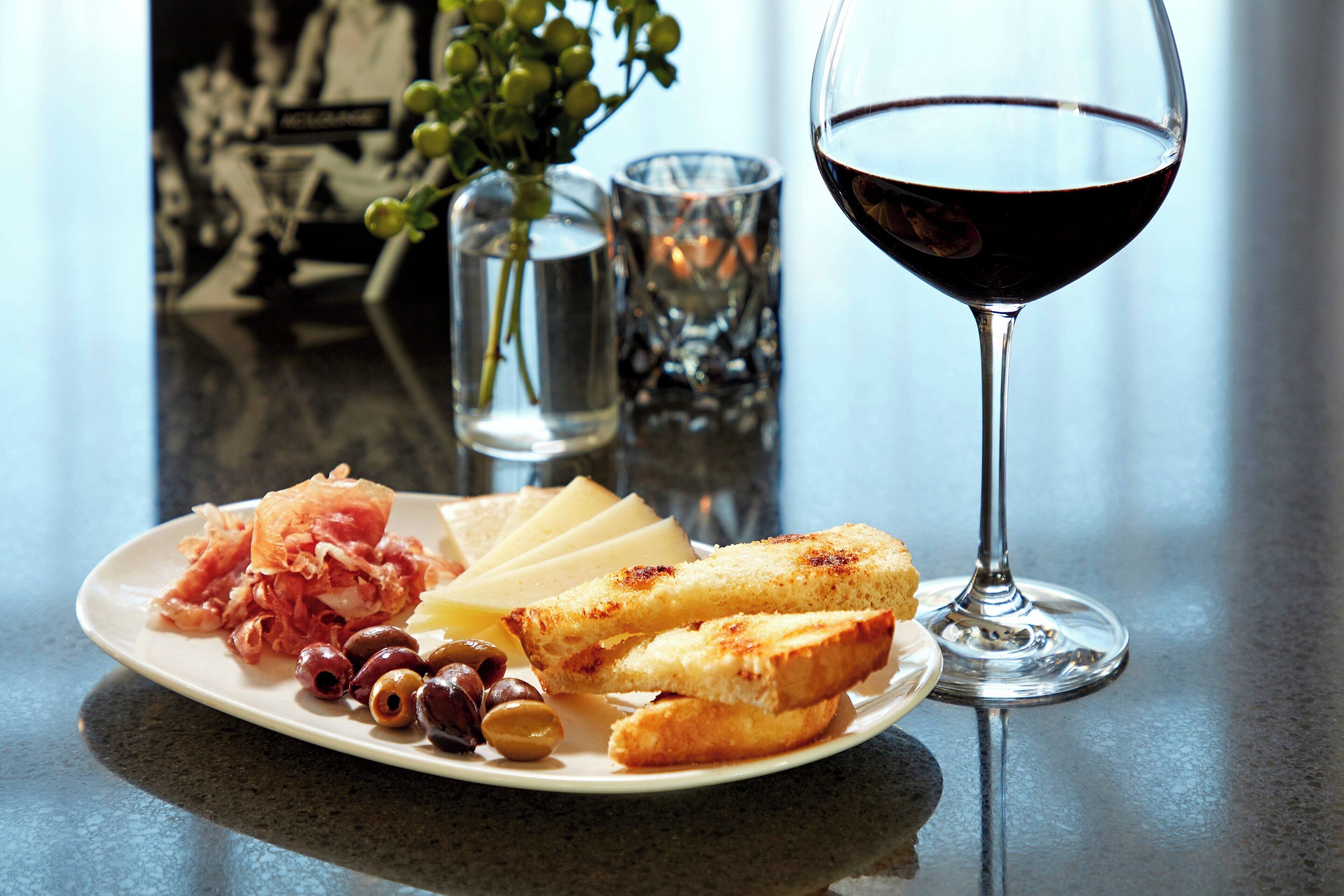 A plate of cheese, olives, and prosciutto with a glass of red wine on a table.