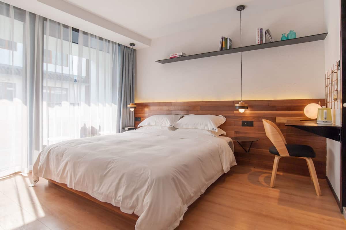A modern hotel room with a large bed, a wooden headboard, a desk with a chair, and a shelf with books and decorative items.