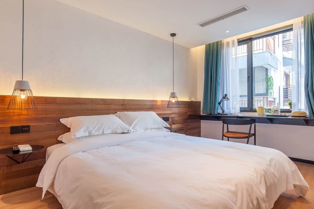 A modern hotel room with a wooden headboard, a large bed with white linens, a nightstand with a lamp, and a view of the city through a large window.