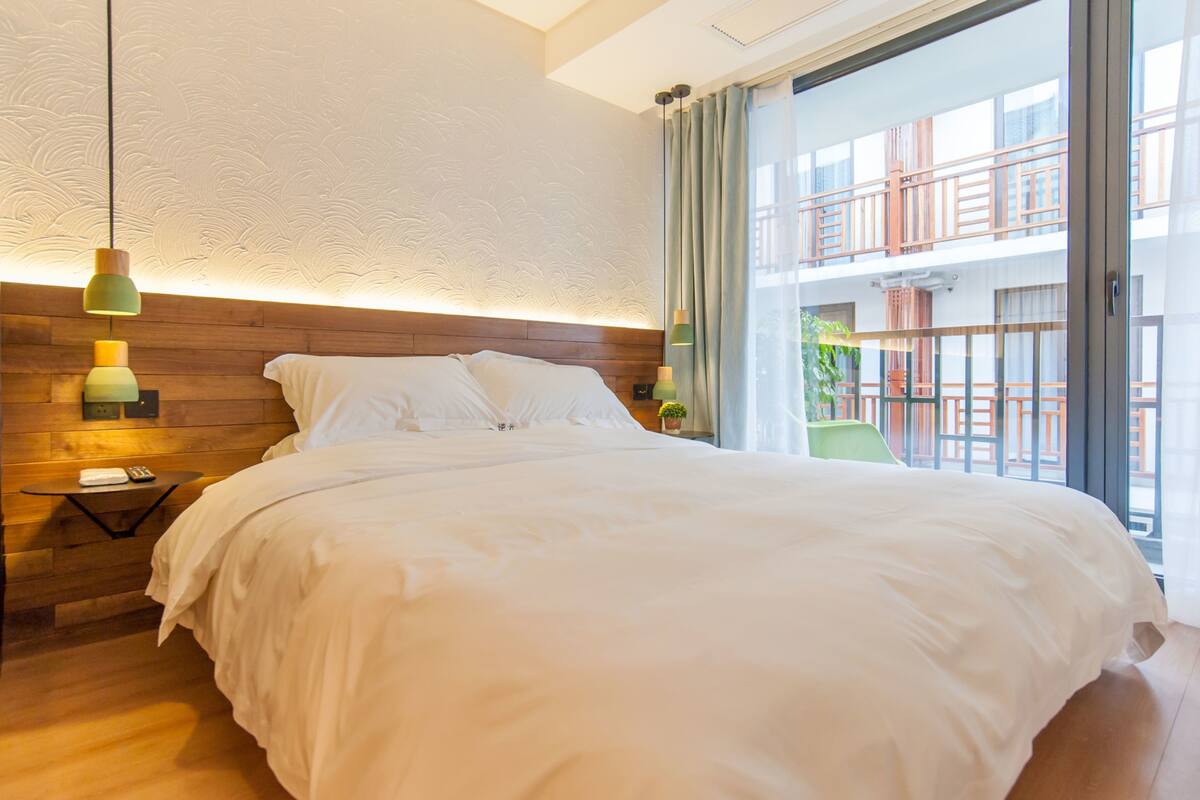 A neatly made bed in a hotel room with a wooden headboard, a nightstand with a phone and remote, and a view of the outside balcony.