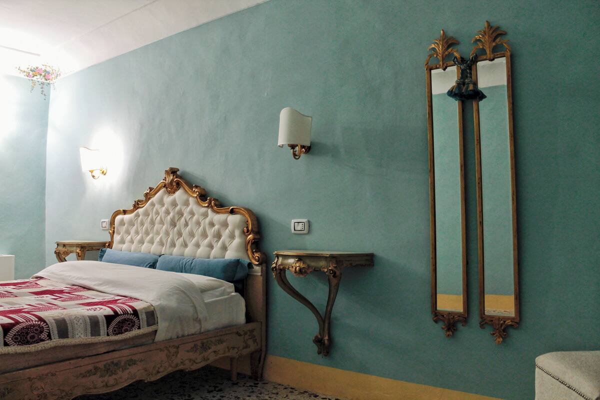 A bedroom with a tufted headboard, a bedside table, a nightstand, a wall-mounted mirror, and a wall sconce.