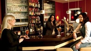 A bar area with shelves of bottles and glasses, a counter, and a person serving a drink. - Hotel 4 Winden (Windhagen)