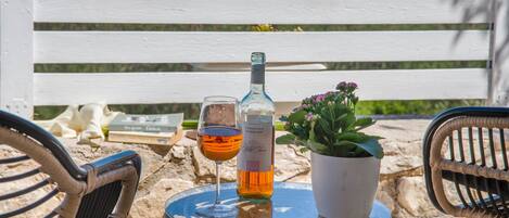 A glass table with a bottle and a glass of wine, a potted plant, and a white bench in the background.