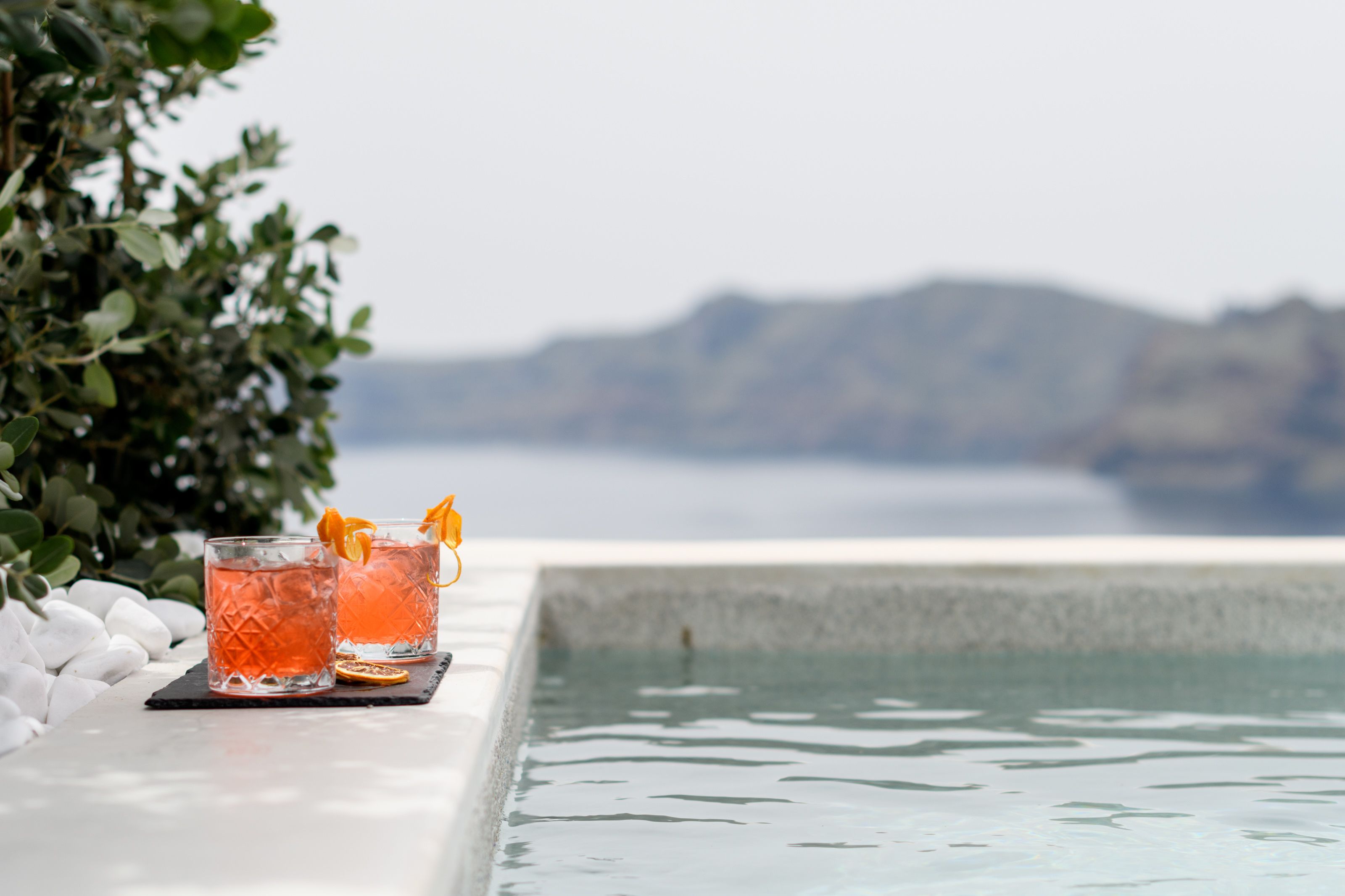 Two glasses of pink drink with orange garnish on a table by a pool with a mountain view.