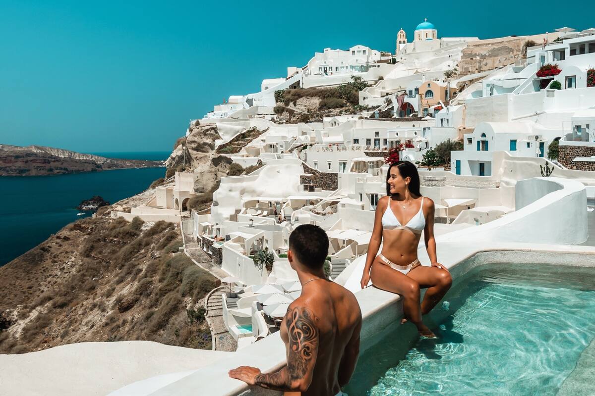 A couple in a white bikini by a pool with a view of white buildings and a blue sea.
