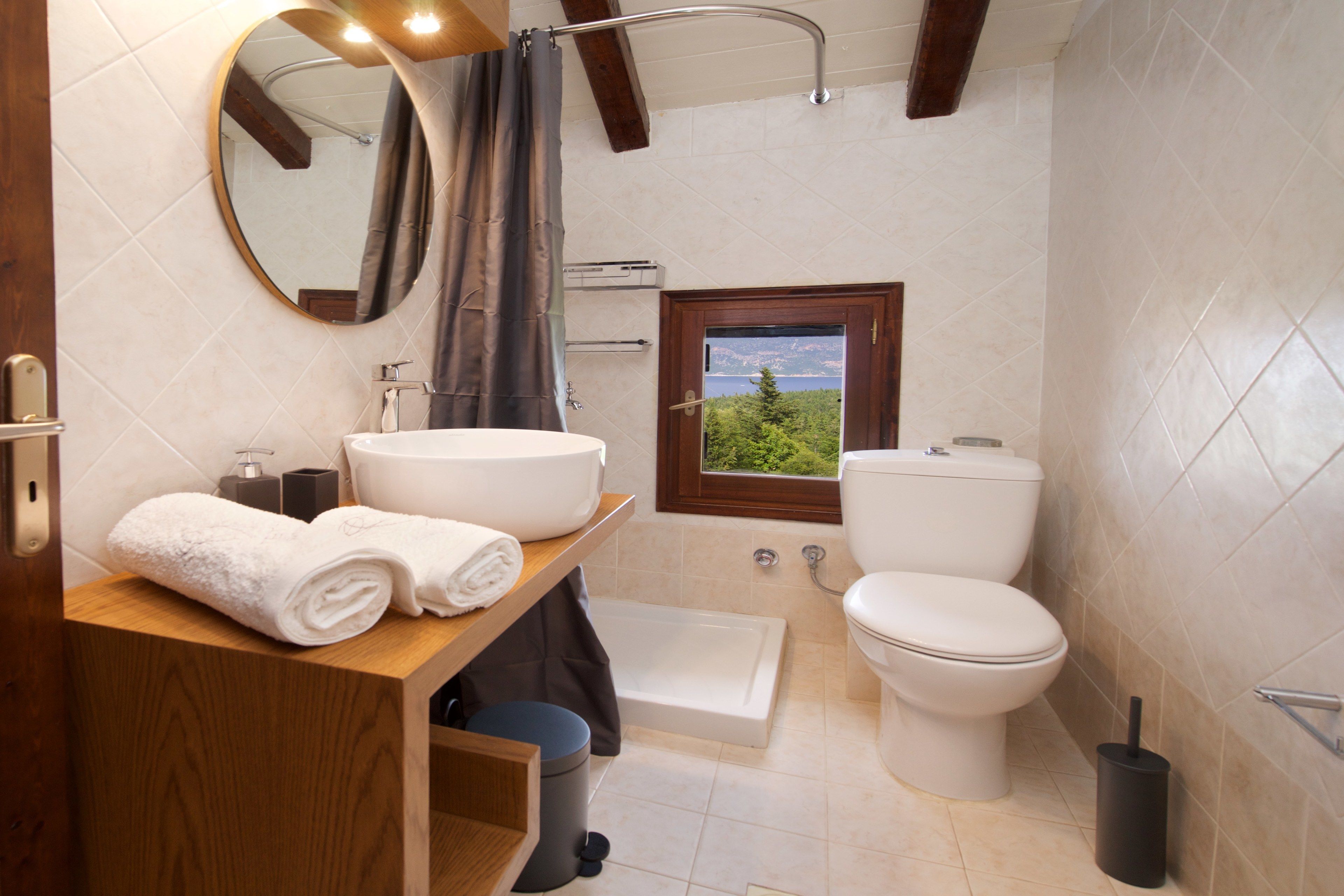 Un baño con tocador de madera, un espejo redondo, una bañera, un inodoro y una ventana con vistas a la vegetación.