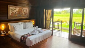 A large bed with white linens, a wooden headboard, and a painting above it. There is a balcony with a view of a green field. - Adil Villa and Resort (Ubud)