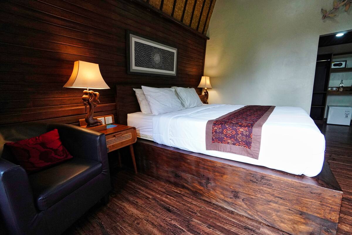 Une chambre d’hôtel avec une tête de lit en bois, un lit recouvert de draps blancs et d’un couvre-lit à motifs, un fauteuil en cuir avec un coussin rouge et deux lampes de chevet.