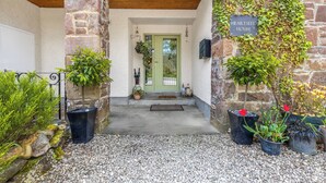 Une entrée en pierre, avec une porte verte, une enseigne indiquant « Heartseed House » et divers pots de plantes.