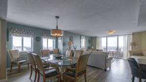A spacious living area with a dining table, chairs, and a sofa. There are large windows providing natural light and a view of the outdoors. - Yacht Club S #1-1001b 2 Bedroom Condo by RedAwning (North Myrtle Beach)