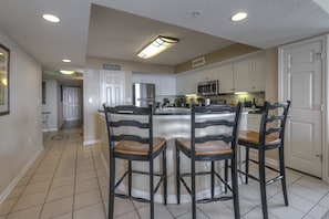 A modern kitchen with white cabinets, a central island with bar stools, and a "Welcome" sign. - Yacht Club S #1-1001 3 Bedroom Condo by RedAwning (North Myrtle Beach)