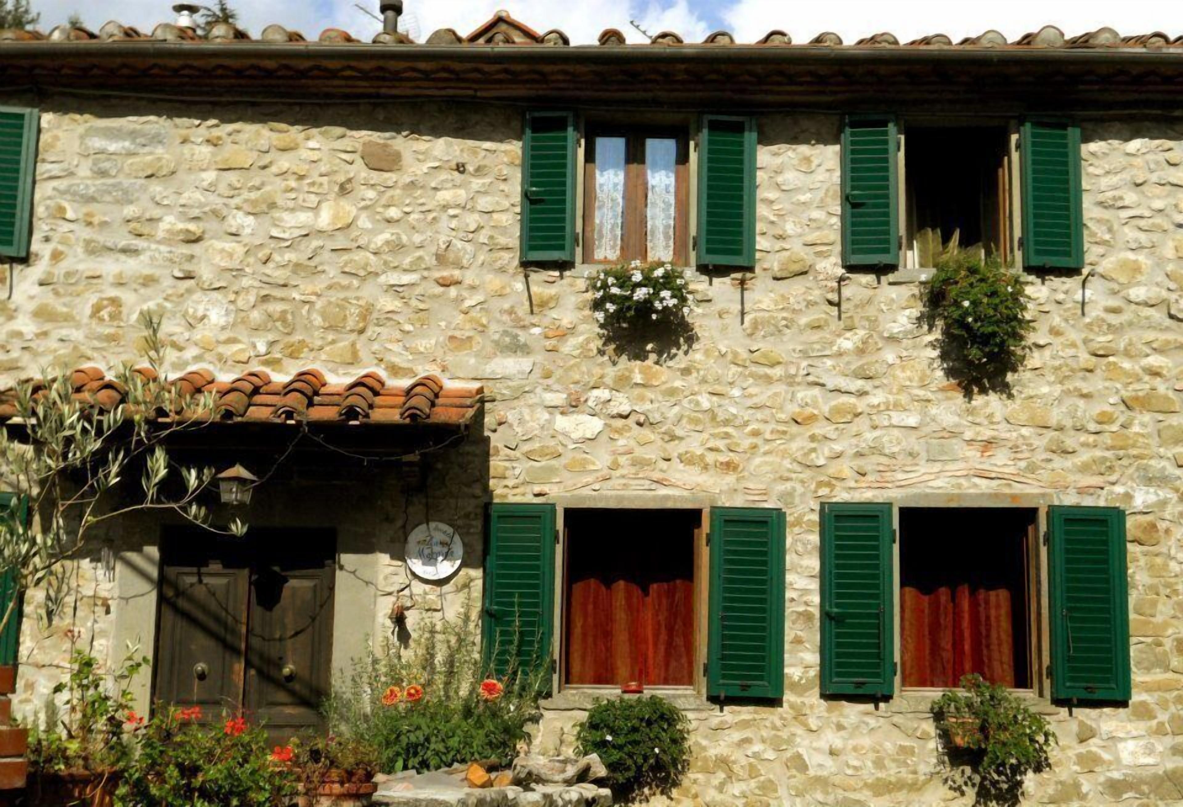 Un edificio de piedra con contraventanas verdes, techo de tejas y un pequeño jardín con flores y plantas.