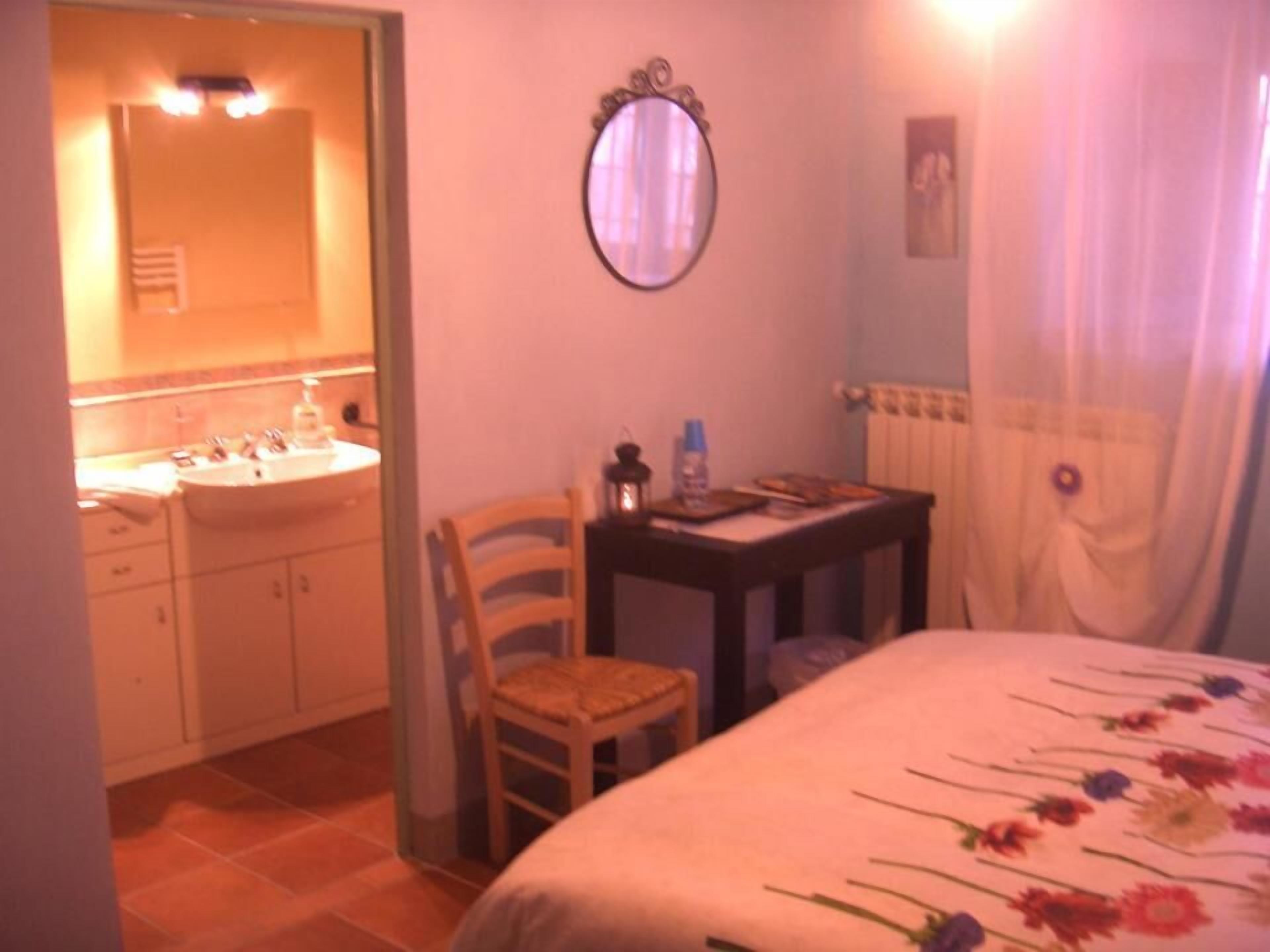 A bedroom with a bed, a chair, a small table with a lamp, a mirror, and a bathroom with a sink and a towel rack.