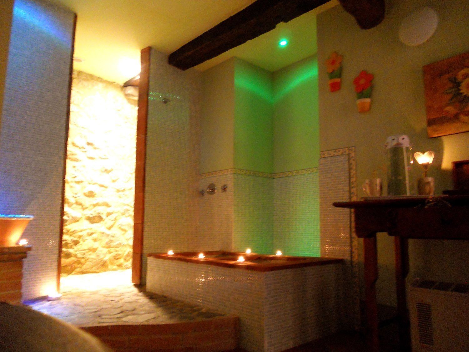 Una stanza scarsamente illuminata con una parete in pietra, un angolo illuminato di verde e candele su un tavolo di legno.