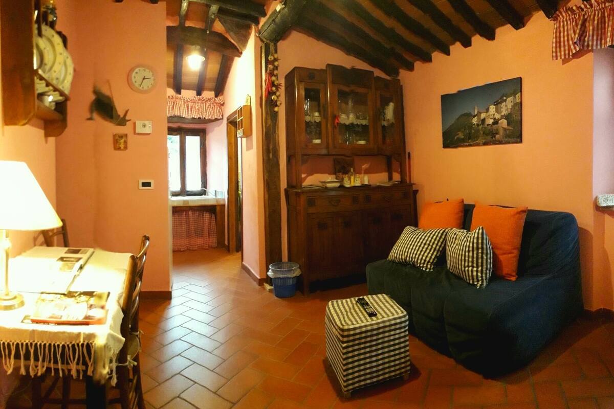 Una sala de estar acogedora con comedor, un sofá con cojines, una mesa con lámpara y una zona de cocina con armarios.