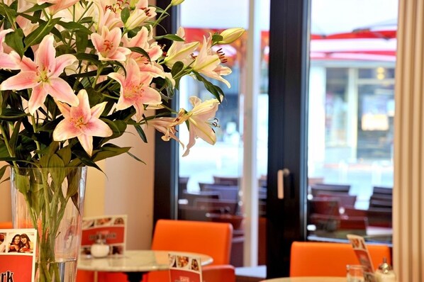 A dining area with orange chairs and a table, a vase of pink lilies, and a window with a view of the outside.