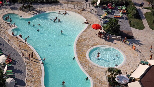 A large, irregularly shaped pool with multiple sections, surrounded by a stone deck and shaded by umbrellas. - Camping Baia Verde (Manerba del Garda)