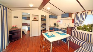 A compact, well-lit room with wooden flooring, a dining table with blue placemats, a striped sofa, and a kitchenette area. - Camping Baia Verde (Manerba del Garda)