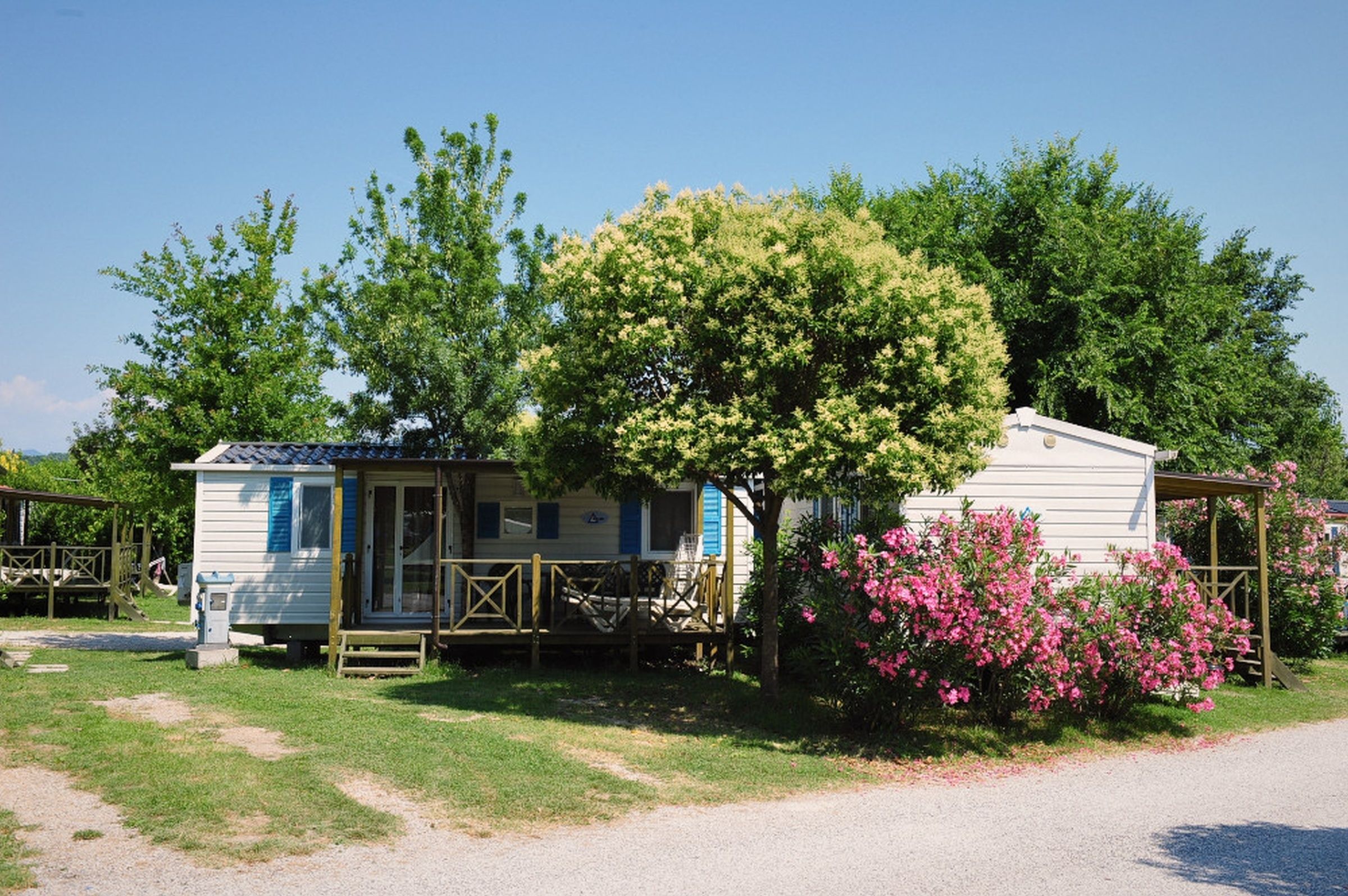 Une maison mobile dotée d’un porche et d’une petite terrasse, entourée d’arbres et d’arbustes en fleurs.
