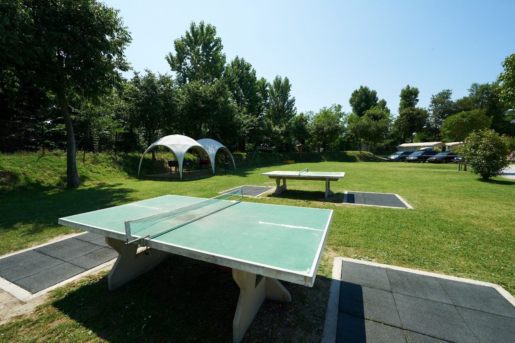 An outdoor table tennis court with two tables and a net, surrounded by grass and trees.