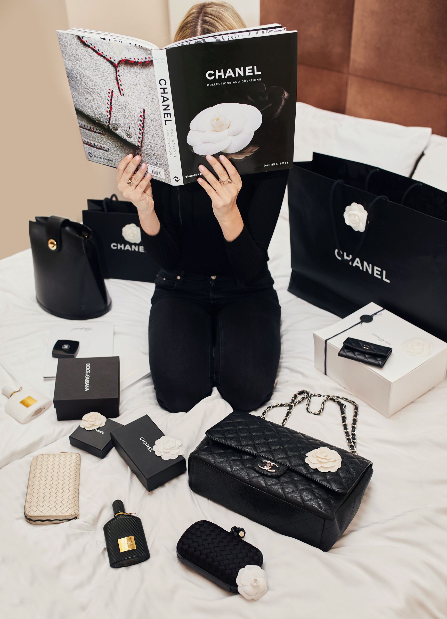 A person sitting on a bed surrounded by various Chanel products including bags, boxes, and a perfume bottle.