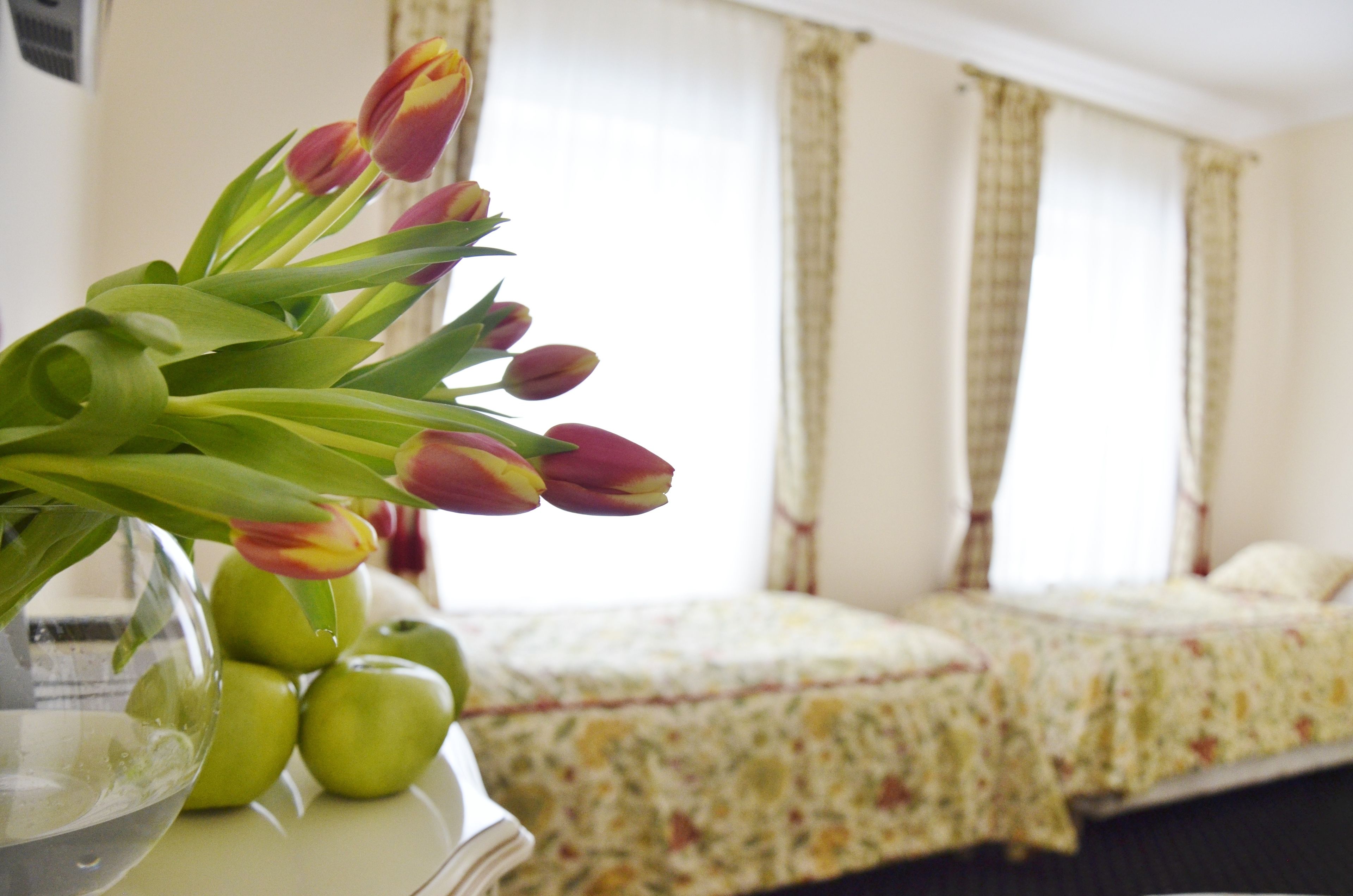 Une chambre avec un lit, une table de chevet ornée d’un vase de tulipes et de pommes, et une fenêtre avec des rideaux.