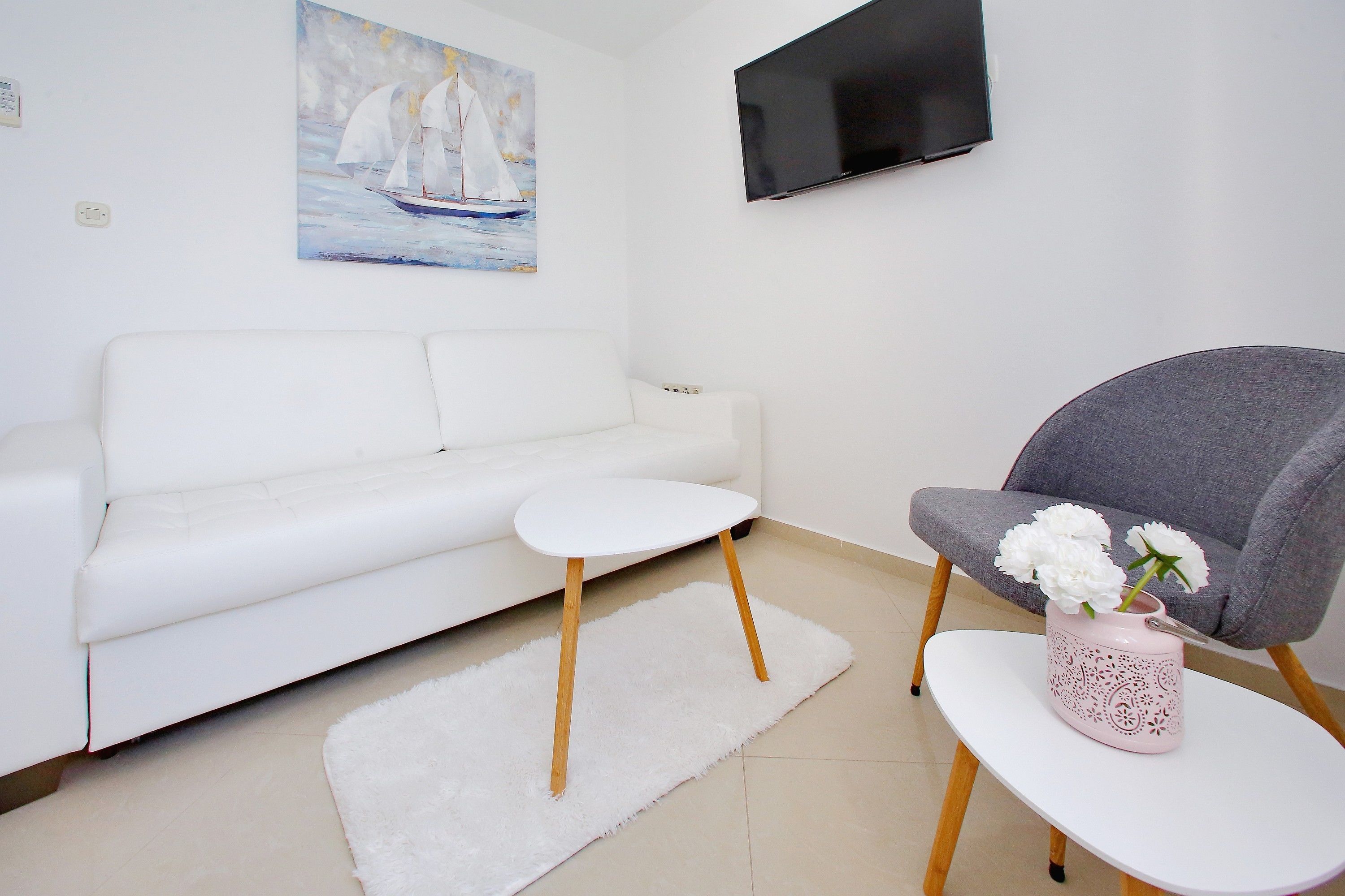 A modern living room with a white sofa, a small white table, a grey armchair, and a pink vase with flowers.