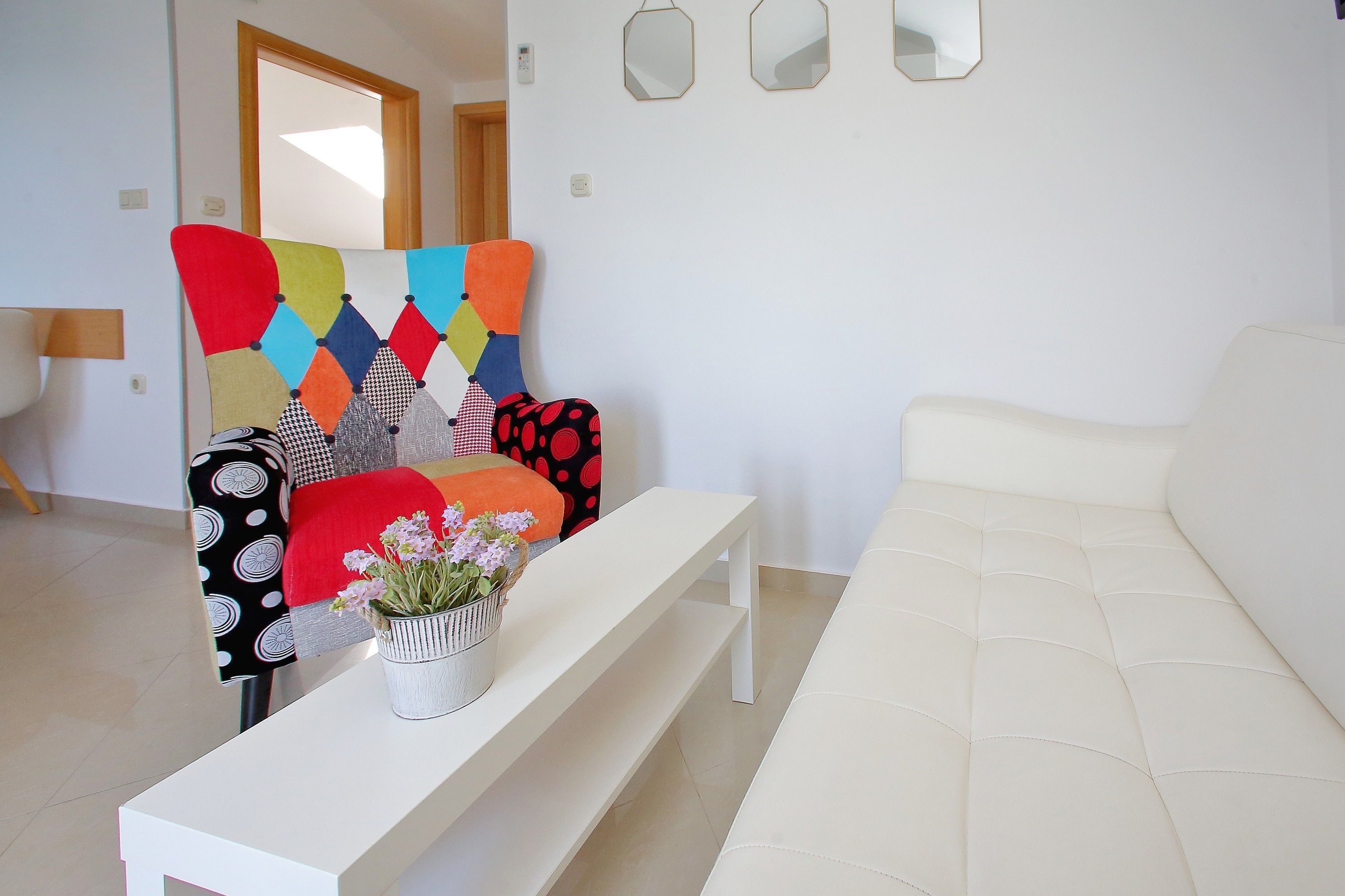 A modern living room with a white sofa, a colorful armchair, a white coffee table with a flower vase, and a white bench.