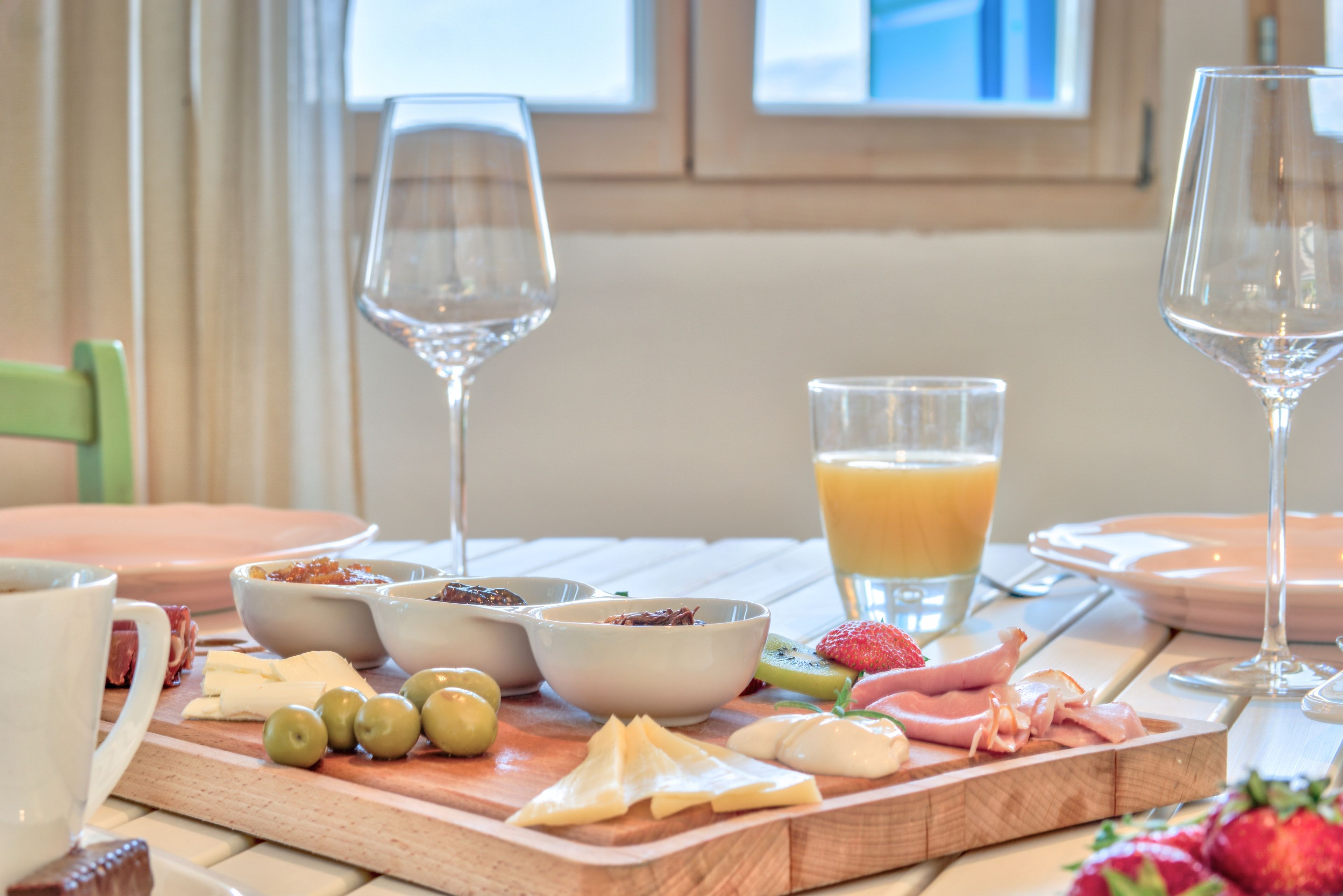 A breakfast setting with a wooden board containing various foods, a glass of orange juice, and two wine glasses.