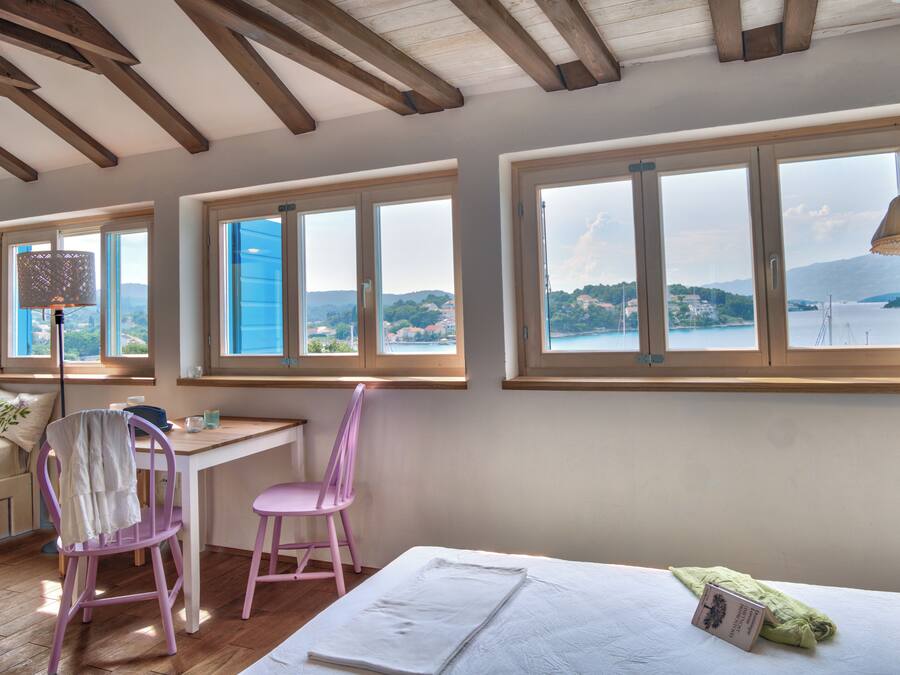 A room with a bed, a desk, and two chairs. There are large windows with a view of a body of water and hills.