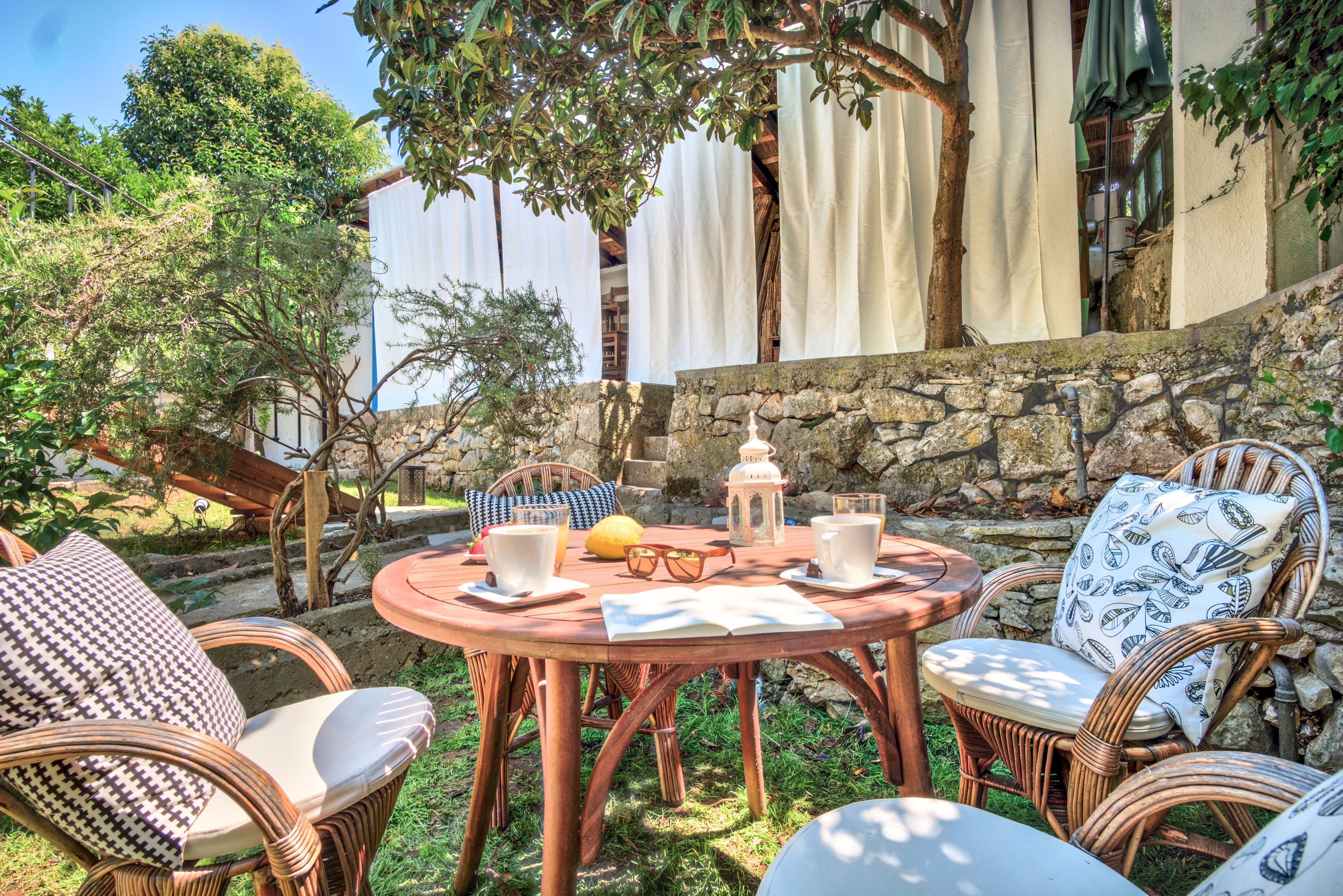 Un espace extérieur aménagé avec des chaises en osier et une table ronde, entouré de verdure et d’un mur en pierre.